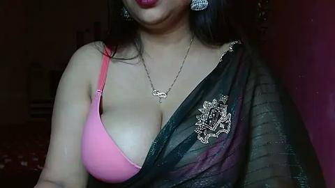 Media: Video of a South Asian woman with medium skin tone, wearing a pink bra and a black sheer saree with floral embroidery. She has long, dark hair and wears silver earrings. The background is dimly lit with red walls.