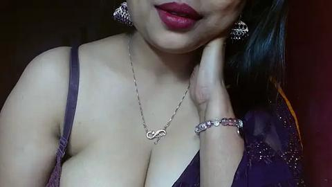 Media: A close-up video of a light-skinned woman with full lips, wearing a low-cut purple dress with a silver necklace, and matching silver earrings. She has her hand on her cheek, with a bracelet on her wrist.