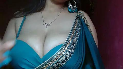 Media: Video of a woman with medium brown skin and long black hair, wearing a revealing turquoise sari with gold embroidery. She has a large cleavage and wears a silver necklace with a pendant. The background is dark red.