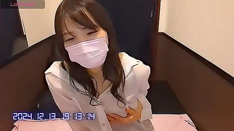 Media: Video of an Asian woman with long black hair, wearing a white shirt, face mask, and glasses, sitting in a dark room, looking down.