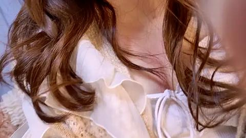 Media: Video of a close-up of a woman's face, focusing on her long, wavy brown hair, wearing a white blouse with lace details, and a beige cardigan. The background is blurred, highlighting her soft features and delicate attire.