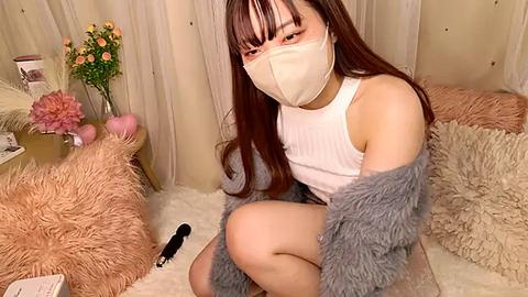 Media: Video of an East Asian woman with long brown hair, wearing a white tank top, gray fur shawl, and a beige face mask, sitting on a plush rug in a cozy, floral-themed room.