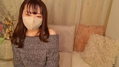 Media: Video of an East Asian woman with long dark hair, wearing a gray off-shoulder sweater, beige mask, and seated on a white couch in a cozy room with beige curtains, a fluffy pillow, and decorative objects.