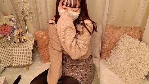 Media: Video of an Asian woman in a beige knit sweater, brown skirt, and face mask, sneezing, seated on a plush, beige carpeted floor. Background includes a floral-patterned table, fluffy pillows, and a tree-shaped decoration.