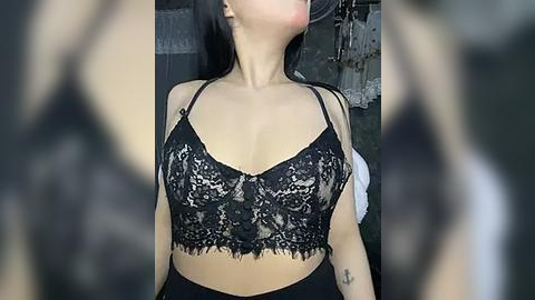 Media: A video of a woman with long black hair, wearing a black lace bralette with thin straps, showcasing medium-sized breasts. The background is blurry, featuring dark, possibly industrial, elements.