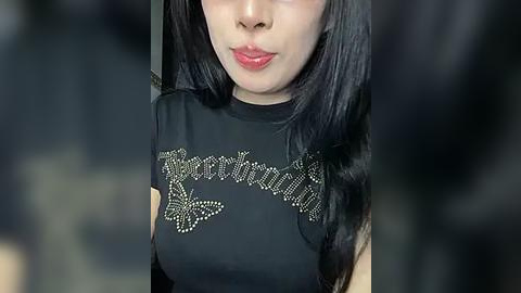 Media: Video of an Asian woman with long black hair, wearing a black top with sequined text \"Lolita\" and a butterfly design. She has fair skin and wears light makeup.