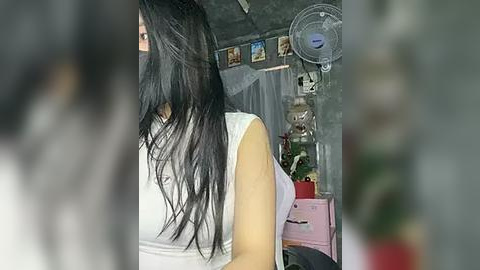 Media: A blurry video of a woman with long, black hair, wearing a white tank top, partially obscured by a person's head. Background features a gray wall with a fan, plants, and a pink box.