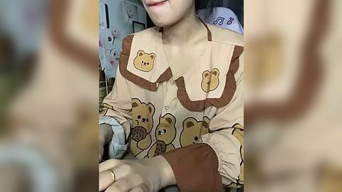 Media: Video of an Asian woman wearing a beige blouse with brown teddy bear patches, sitting indoors with blurred background.