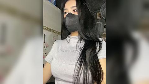 Media: A video of a young woman with long black hair and fair skin, wearing a gray t-shirt, black face mask, and a black headband, indoors with blurred background.