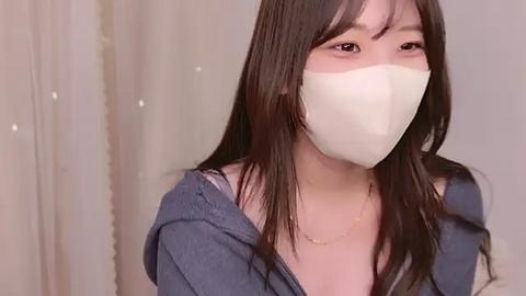 Media: Video of an East Asian woman with long black hair, wearing a white surgical mask, a blue hoodie, and a gold necklace, against a backdrop of white curtains.