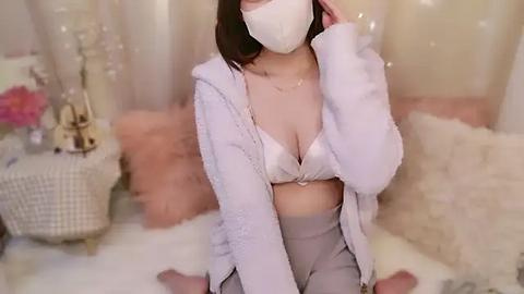 Media: Video of a light-skinned Asian woman with straight dark hair, wearing a white face mask, light pink bra, and fluffy robe, sitting on a fluffy white rug in a cozy, softly lit room.