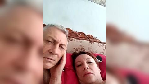 Media: A video of an elderly white couple lying on a red bed with floral-patterned headboard, both appearing tired and relaxed, their faces out of focus.