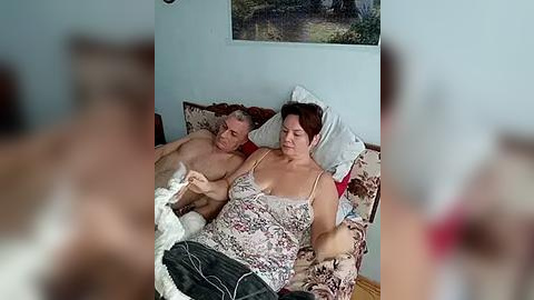 Media: Video of an elderly couple in bed, with the man shirtless and frail, and the woman in a floral nightgown, surrounded by disarrayed bedding and a blurred background.