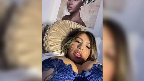 Media: Video of a young Asian woman with light brown skin, long brown hair, and a blue lace bra, lying on a beige bed. A nude painting of a woman's face is visible on the wall.