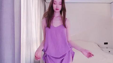 Media: A video of a slender, fair-skinned woman with long brown hair, wearing a loose, light purple nightgown, standing in a modern, minimalistic bedroom with white bedding and curtains.