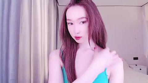 Media: Video of a young, slender, fair-skinned woman with long, straight, auburn hair, wearing a turquoise bra, standing in a minimalist, softly lit bedroom with white curtains and a bed.