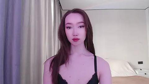 Media: A video of a young, fair-skinned Asian woman with long, straight brown hair, wearing black lace lingerie, standing in a modern bedroom with white walls, a bed, and beige curtains.