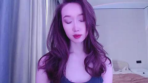 Media: A video of a fair-skinned woman with long, wavy, dark brown hair, wearing a dark blue sleeveless top, standing in a softly lit bedroom with light-colored walls and a bed visible in the background.