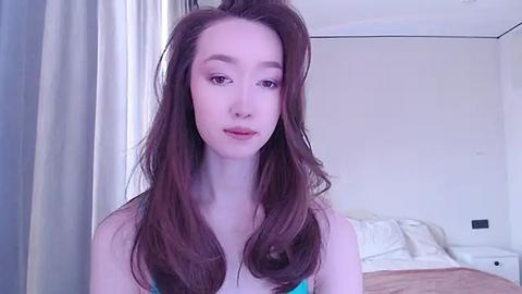 Media: Video of a young woman with long brown hair, fair skin, wearing a blue tank top, standing in a softly lit bedroom with white walls, curtains, and a bed.