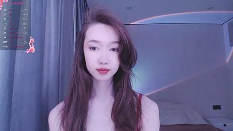 Media: A video of a young Asian woman with long, straight brown hair, fair skin, and red lipstick, wearing a red top. The background shows a bed with white sheets and a large window with gray curtains.