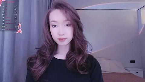 Media: Video of a young Asian woman with long, wavy brown hair, wearing a black shirt, standing in a bedroom with white walls and a bed.