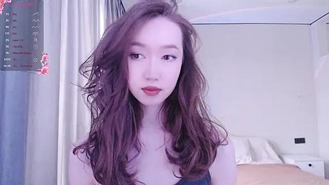 Media: Video of a fair-skinned, young Asian woman with wavy brown hair, wearing red lipstick, standing in a softly lit bedroom with a bed and floral curtains in the background.