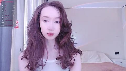 Media: A video of a fair-skinned young woman with long, wavy brown hair, wearing a white tank top, standing in a minimalist bedroom with a white bed and floral curtains.