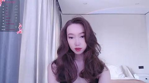 Media: A video of an Asian woman with long, wavy brown hair, fair skin, and a petite build, wearing a black top, standing in a modern, minimally decorated bedroom with white walls, a bed, and a calendar on the left.