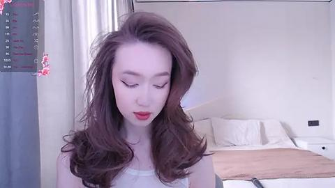 Media: Video of a young Asian woman with fair skin and long, wavy brown hair, wearing a white tank top, standing in a minimalist bedroom with a neatly made bed and white curtains.