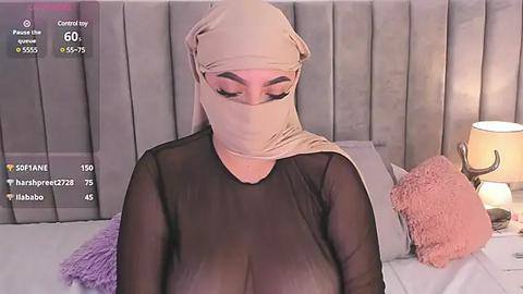 Media: A video of a woman with a large bust, wearing a sheer black top and beige hijab, sitting on a bed with gray headboard and pillows.