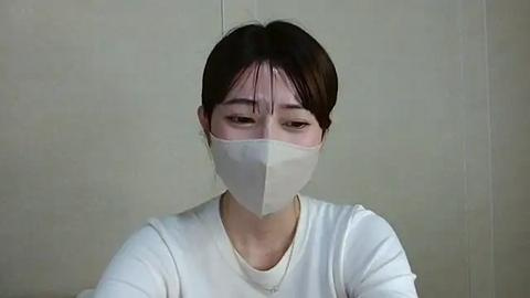 Media: Video of an East Asian woman with dark hair in a bun, wearing a white face mask and plain white top, against a plain beige tiled wall background.