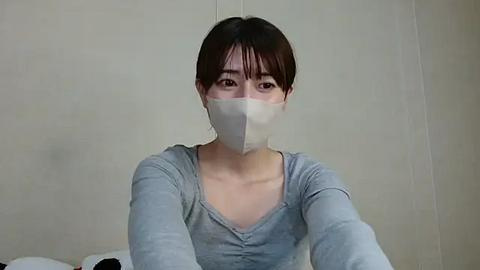 Media: Video of a young East Asian woman with short dark hair, wearing a light gray V-neck shirt and a white surgical mask, sitting in a plain beige room.