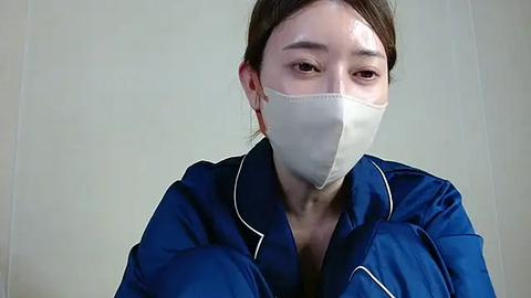 Media: Video of an Asian woman with light skin, brown hair, wearing a blue hospital gown and a white surgical mask, against a plain, light-colored background.