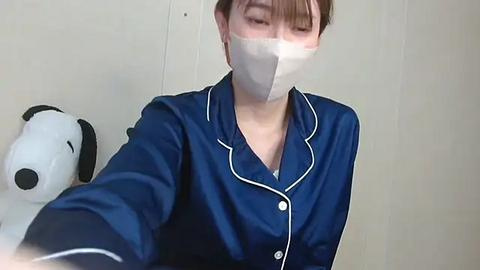 Media: A video of an Asian woman in a blue satin pajama set with white piping, wearing a white surgical mask, and a plush dog toy on a white wall.