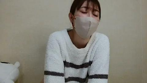 Media: Video of an Asian woman with short brown hair, wearing a white and gray striped towel and a white face mask, leaning forward with eyes closed, against a beige wall.