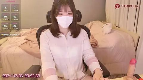 Media: Video of a young Asian woman with shoulder-length black hair, wearing a white face mask and white hoodie, sitting on a black gaming chair, surrounded by a cluttered, beige bedroom.