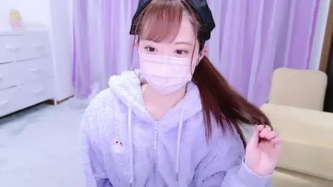 Media: Video of an Asian woman with light skin, straight brown hair, and bangs, wearing a white face mask, black headband, and a fluffy lavender hoodie. She sits on a light gray floor in a room with white furniture and lavender curtains.