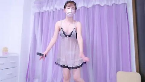 Media: Video of an Asian woman in a see-through maid outfit, holding a remote, in a room with lavender curtains and a white dresser.