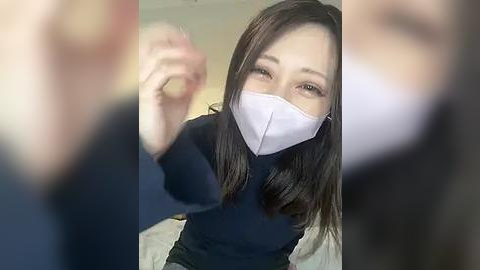 Media: Video of a young East Asian woman with long black hair, wearing a white mask, black top, and holding a coin, blurred background, indoor setting.