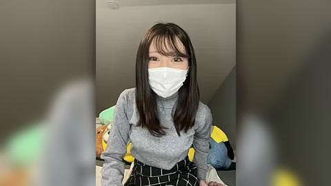 Media: Video of a young Asian woman with long black hair, wearing a white face mask, gray sweater, and black skirt, sitting on a bed with plush toys in a dimly lit room.