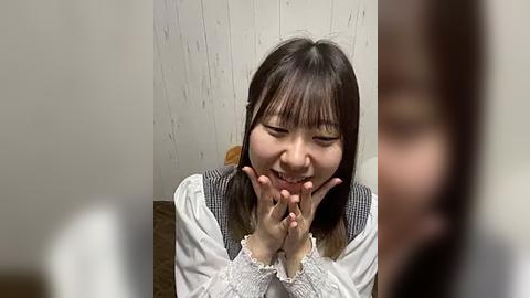 Media: Video of a smiling Asian woman with straight, shoulder-length black hair, wearing a white blouse and gray vest, seated indoors with a white wooden wall in the background.