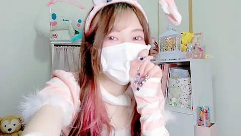 Media: Video of an Asian woman in a pink bunny costume, wearing a face mask, with long, straight, reddish-brown hair, holding a lollipop, in a brightly lit, pastel-colored room.