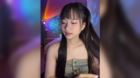 Media: A video of an East Asian woman with long black hair in pigtails, wearing a denim top, sitting in a dimly lit room with colorful neon lights in the background.