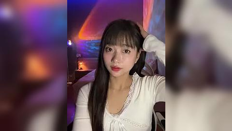 Media: A video of a young Asian woman with long black hair and fair skin, wearing a white lace top, seated indoors with dim lighting and blurred figures in the background.