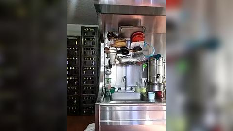 Media: Video of a stainless steel industrial kitchen sink with multiple faucets, surrounded by a cluttered countertop with tools, utensils, and a hanging red and white cloth. Background features a metal shelving unit with various items.