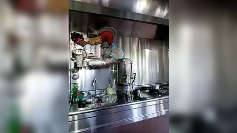 Media: Video of a commercial kitchen with a stainless steel countertop and sink, cluttered with various utensils, pots, and a large, metallic steamer. The background features a shiny, metallic ceiling and walls, creating a sleek, industrial ambiance.