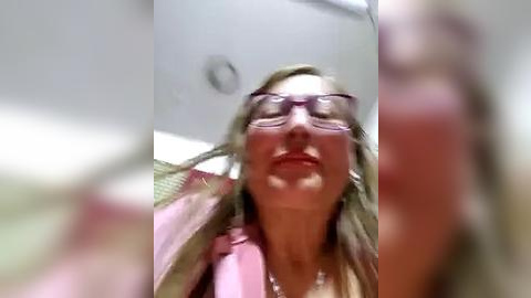 Media: Video of a blonde woman with fair skin, wearing purple glasses and a pink blazer, her mouth agape, in a blurry, out-of-focus indoor setting.