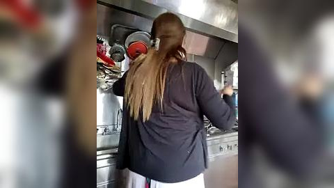 Media: A blurry video of a person with long, straight, blonde hair, wearing a dark jacket, standing in a professional kitchen with stainless steel appliances and utensils on shelves.