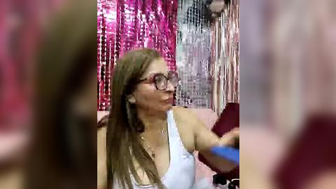 Media: Video of a woman with long, light brown hair, wearing glasses, a white tank top, and a necklace, against a pink and silver tinsel backdrop.