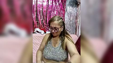 Media: Video of a middle-aged woman with braided hair and glasses, wearing a gray sports bra, seated on a bed. Background features a pink, sequined curtain and a red wall.